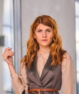The image portrays a red-haired, professionally dressed woman in her mid-thirties, standing in what appears to be an office building. 