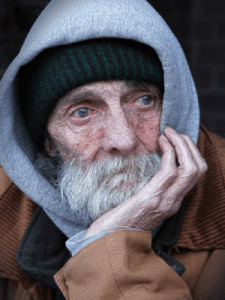 The image portrays an elderly man, dressed in a jacket, hoodie, and winter cap, staring off into the distance. 