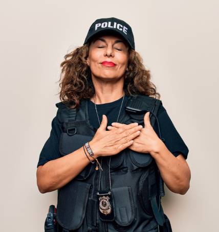 Image depicts a middle age policewoman wearing bulletproof uniform and cap over white background smiling with hands on chest, eyes closed with grateful and kind expression.