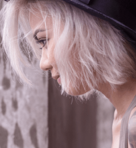 The image features a white woman in her early twenties who has bleached-blond hair and a tattoo that extends from her shoulder to her arm. Completing her edgy look, she wears a black fedora hat.