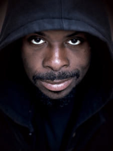 The image depicts a young black man, wearing a dark hoodie, with a slight smile, looking directly into the camera.
